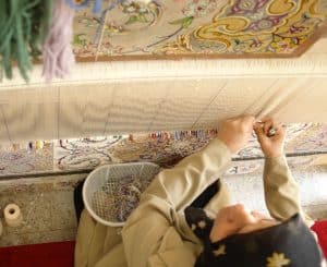 A woman focused on her artwork, using brushes and colors to create a Persian rug masterpiece.